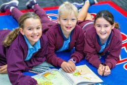 Three students lay on a bright blue and red matt in a classroom. The students have a picture book open on the ground in front of them.